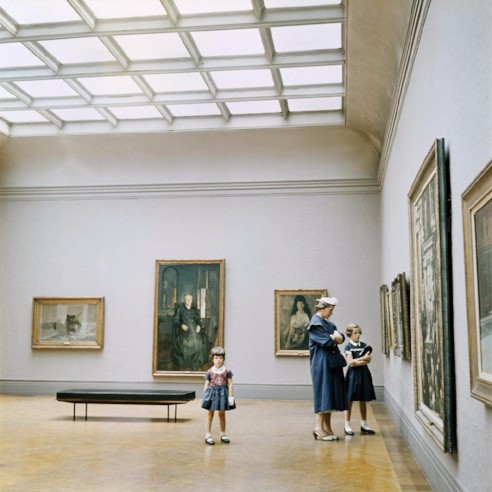Photo of a mother with two daughters at an art gallery, dressed in 1940s clothing. The mother and eldest daughter stand in front of a gigantic painting with their arms crossed, looking stern. The youngest daughter stand apart in the centre of the room looking directly at the camera.