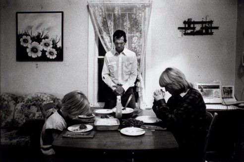 Black and white photo of a family at a table saying Grace before diner. A mother and child sit opposite each other with heads bowed. A father stands between them at a window, eyes closed. A picture of sunflowers is on the wall to one side, above a floral sofa.