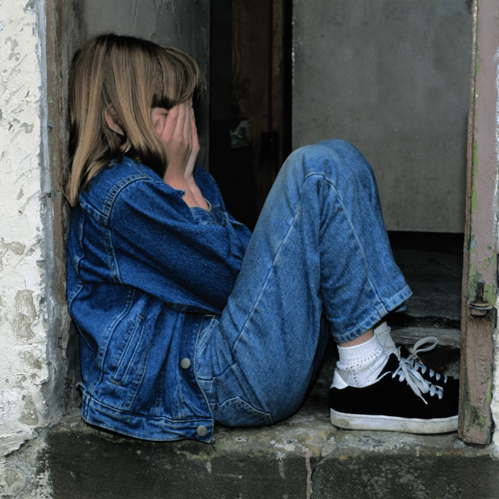 A child wearing jeans and a denim jacket sitting in a doorway with her hands covering her face. 