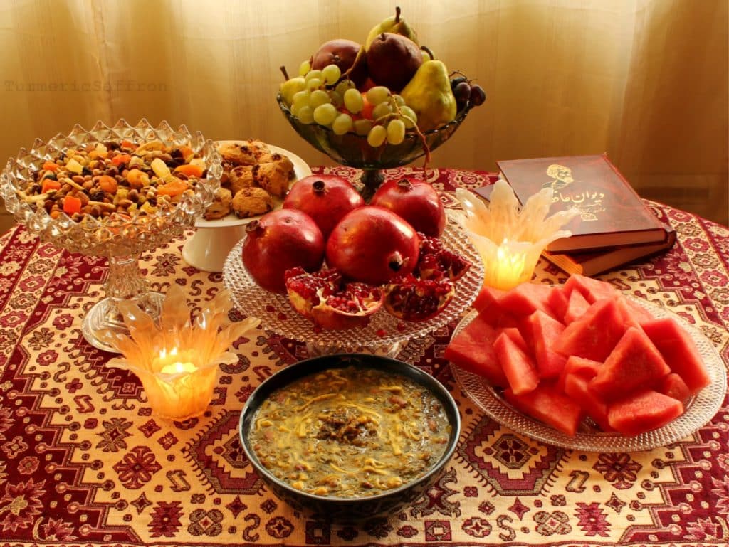 Photograph of a Yalda spread on a patterned tablecloth: bowls of fruit, nuts, pomegranates, watermelon and cakes, along with candles and books of poetry. 