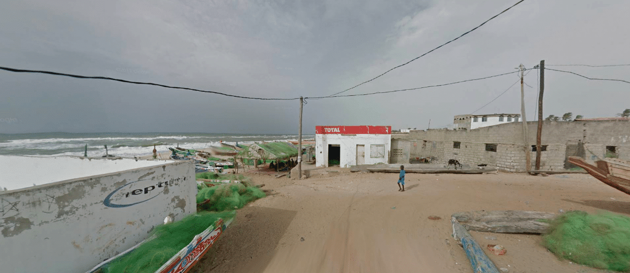 Google Earth street view image of a beachside area in Thiès, Senegal. On there is a short beach full of small fishing boats piled with green nets. Slightly futher inland there are some single storey flat roofed buildings and a sandy flat street. A small figure in blue stands in the centre. Electricity poles and wires stretch overhead and the sky looks grey and cloudy. 