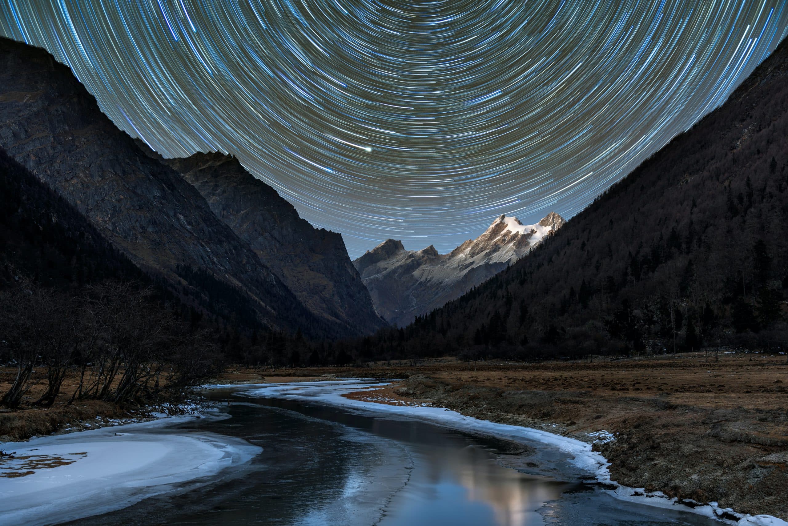 A timelapse photo of mountains above an icy river that shows the movement of the stars in a series of spiraling lights