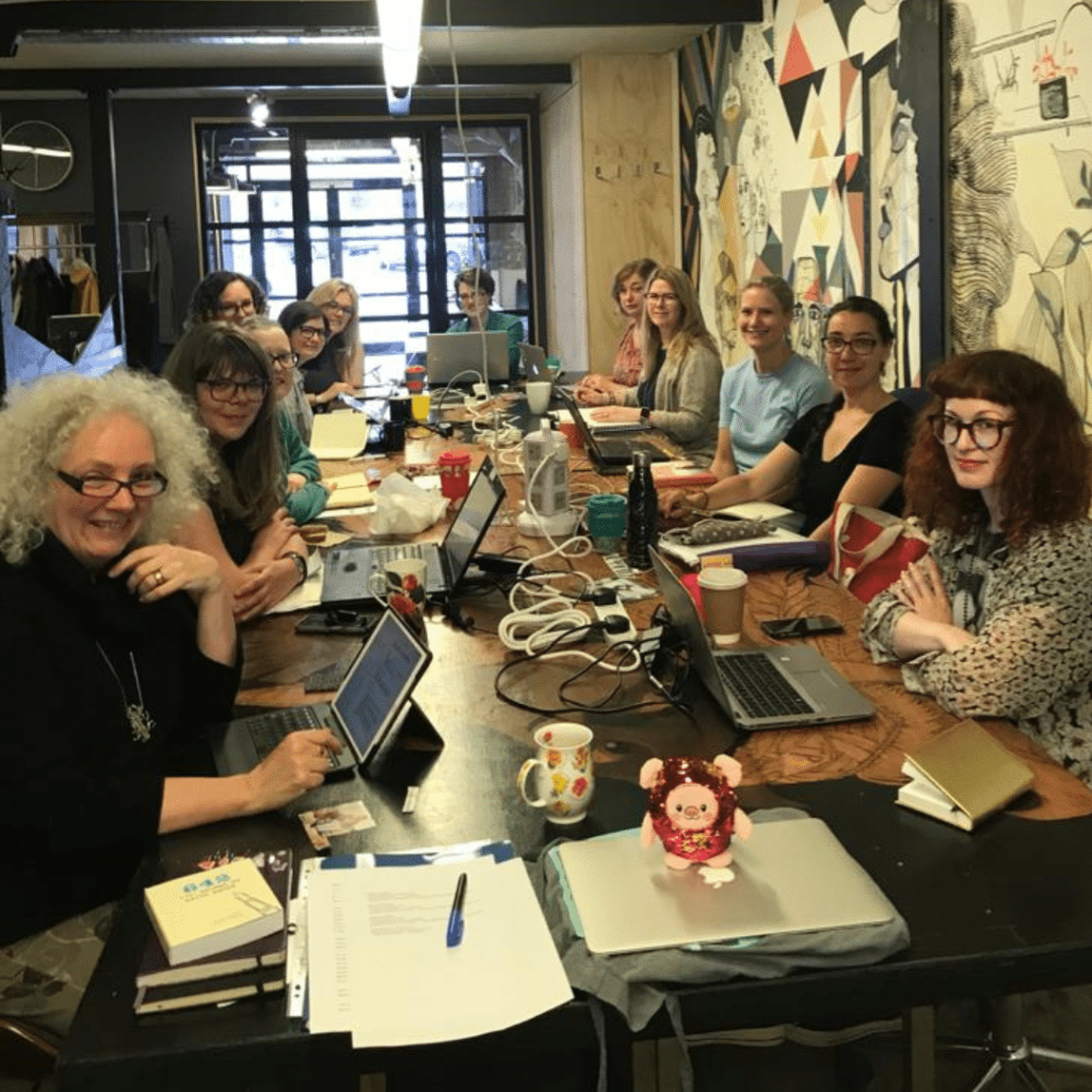 A photo from one of our Edinburgh writing retreats showing a table full of smiling writers with laptops and notebooks and our special mascot, Glitter Pig. 